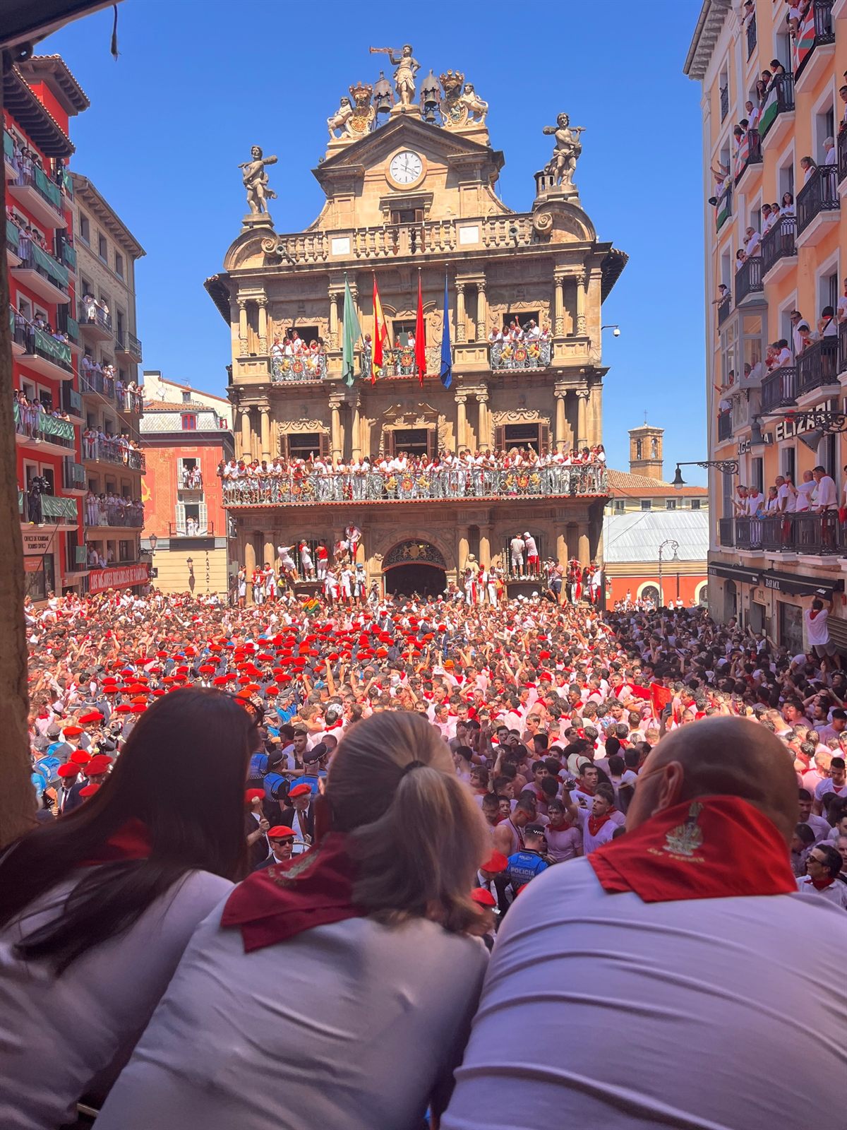 Tres amigas con pañuelo rojo miran desde un balcón la plaza del Ayuntamiento llena durante el Chupinazo